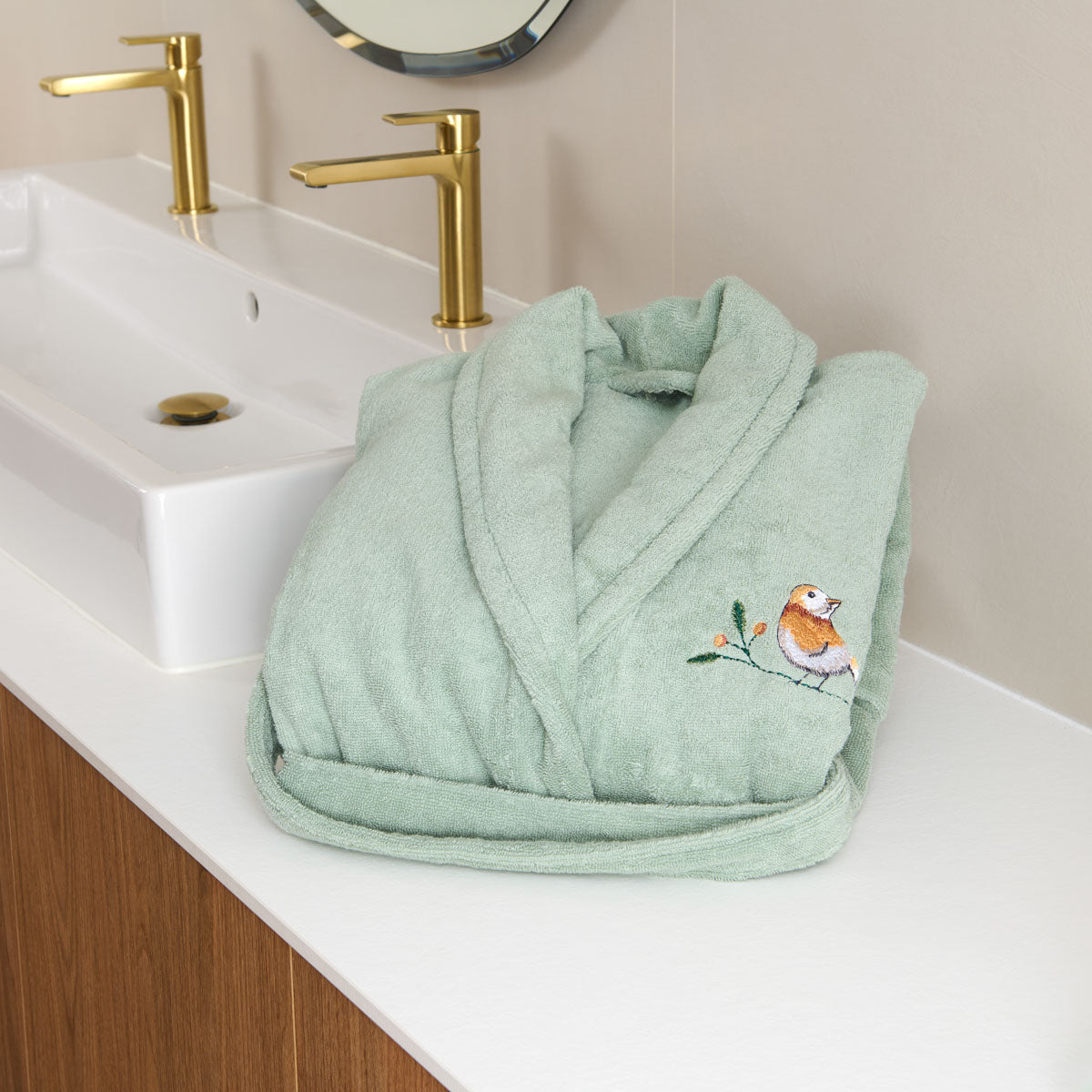 Peignoir femme Doux Refuge en coton éponge Céladon, plié dans une salle de bain avec broderie oiseau apparente